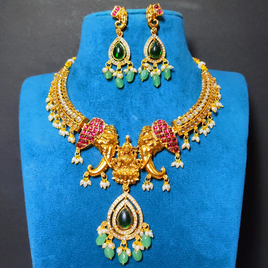 Antique Gold Finish Indian traditional jewelry set featuring a necklace which has Goddess Lakshmi flanked with twin elephants and matching earrings with green teardrop stones, white pearl-like accents, displayed on a blue velvet stand.