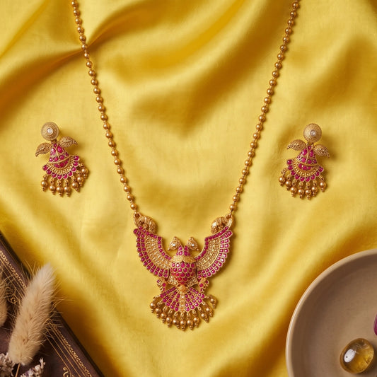 A traditional South Indian Gandaberunda temple jewelry set featuring a matte antique gold toned pendant necklace and matching earrings. The intricate pendant depicts the mythical royal two-headed bird adorned with radiant ruby-red kemp stones and delicate dangling gold toned beads, suspended from a gold toned bead chain.