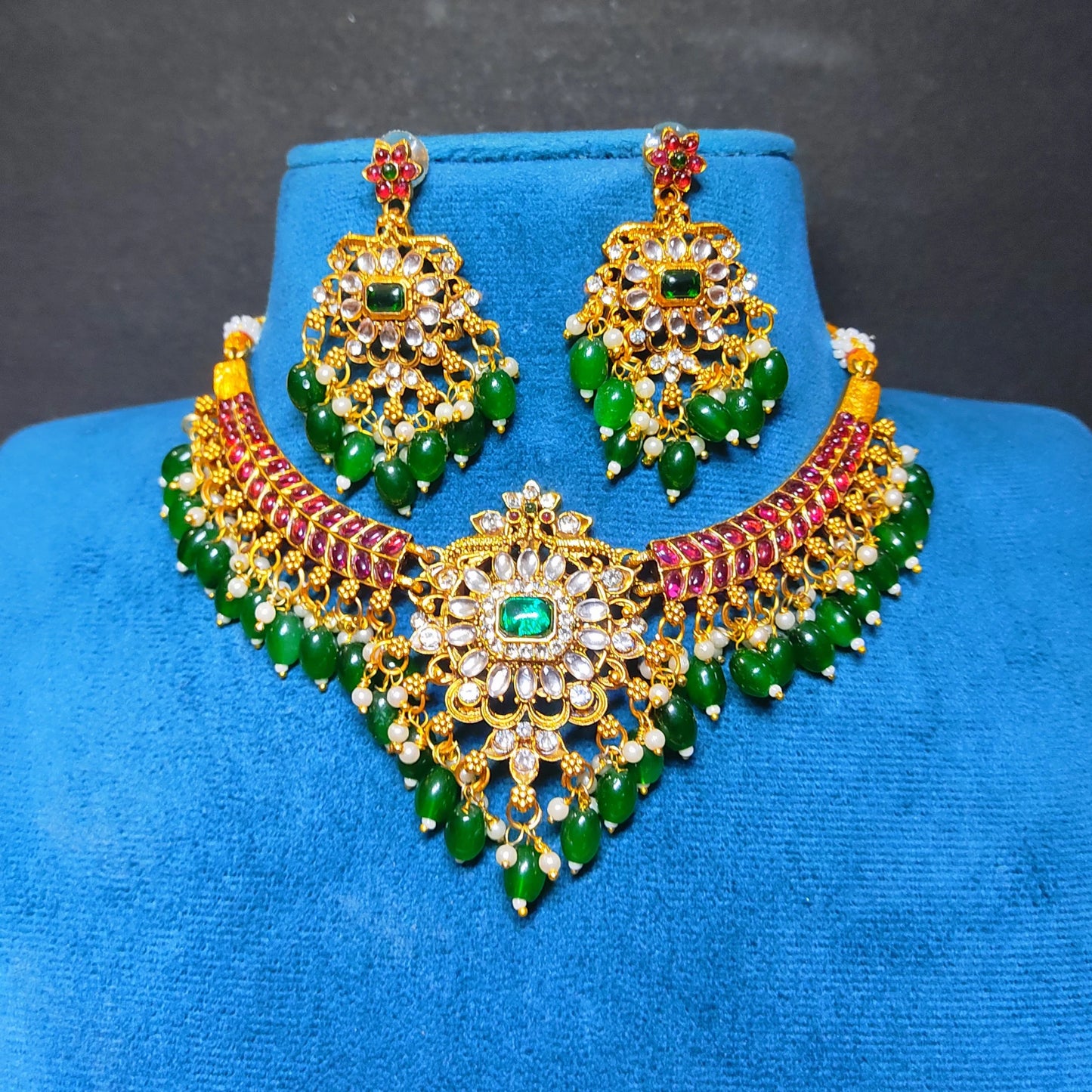 An ornate Indian kundan jewelry set featuring a gold-toned necklace and matching earrings, embellished with green stones, red stones, and clear crystals, displayed on a blue velvet stand.