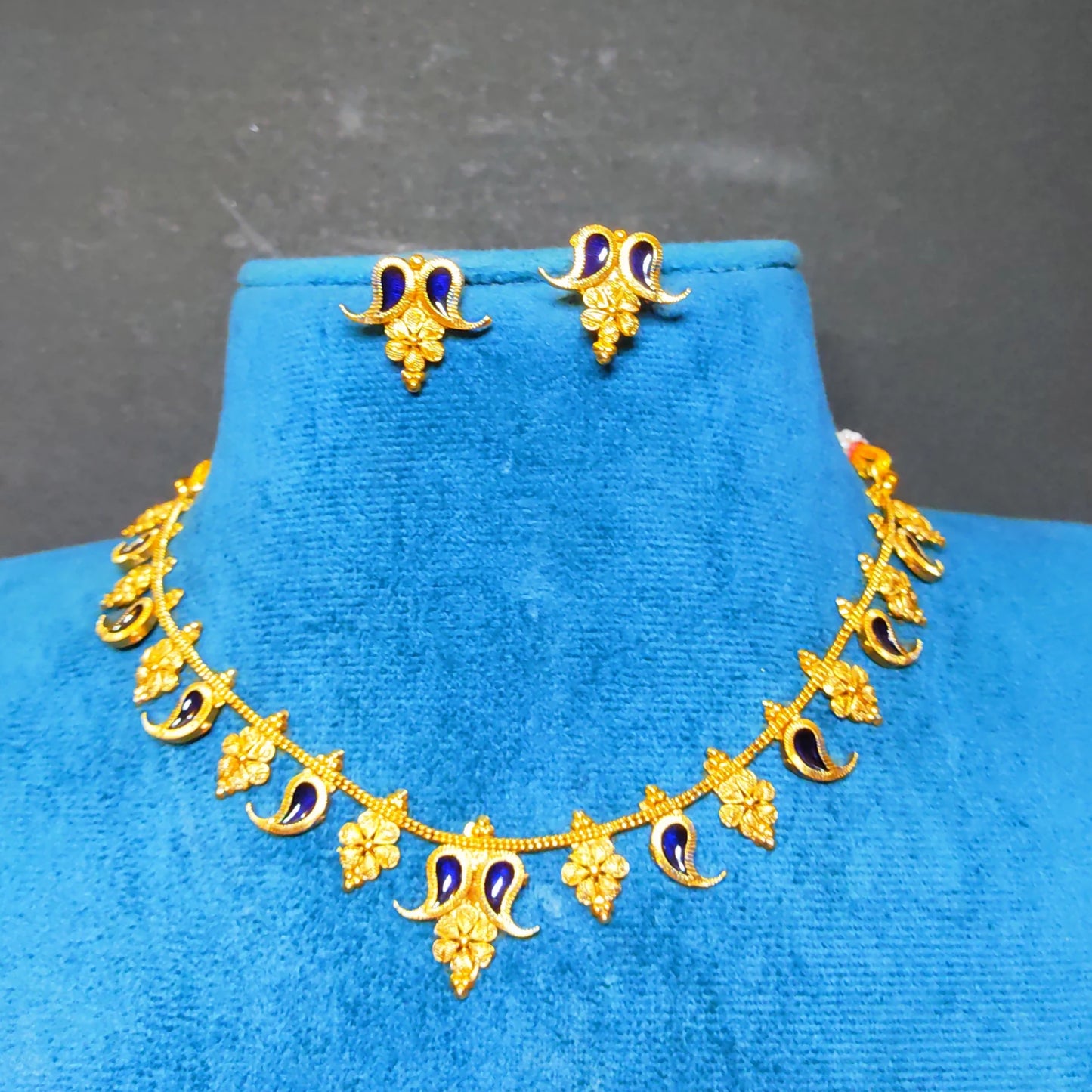 Gold-toned jewelry set including a necklace and matching earrings with blue enamel accents, displayed on a bright blue velvet jewelry bust against a dark gray background.