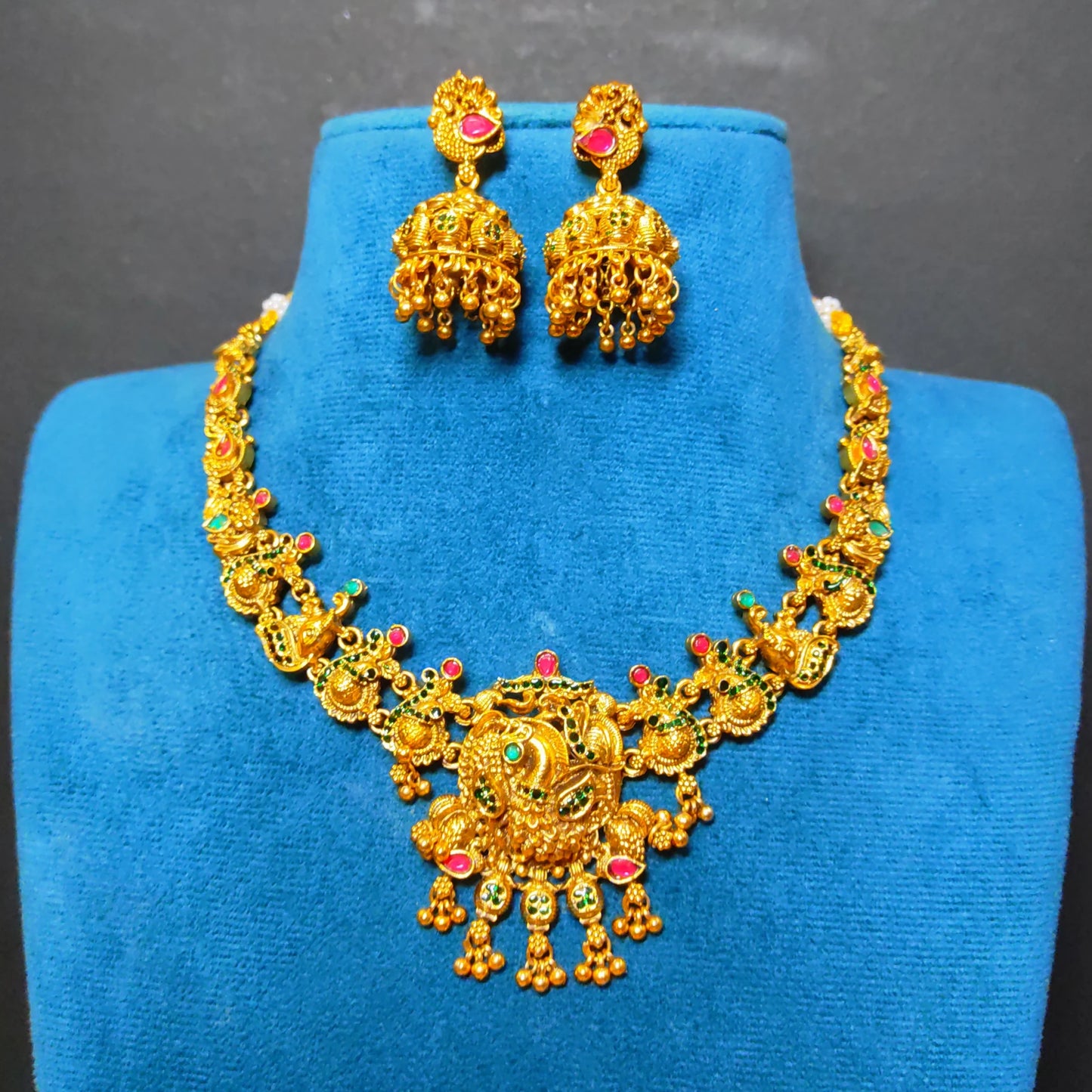 Antique Gold-toned traditional jewelry set featuring a necklace and matching earrings displayed on a blue velvet jewelry bust. The pieces are decorated with red and green stone accents and has peacock motif as the central pendant