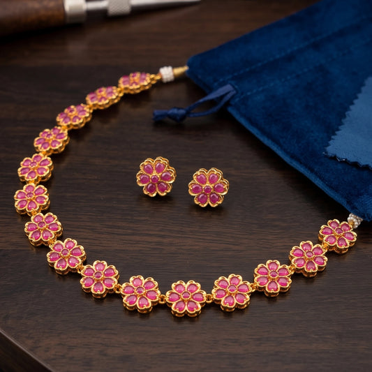 Antique Gold finish floral necklace and matching stud earrings with pink accents, displayed on a dark wooden surface next to a blue velvet pouch.