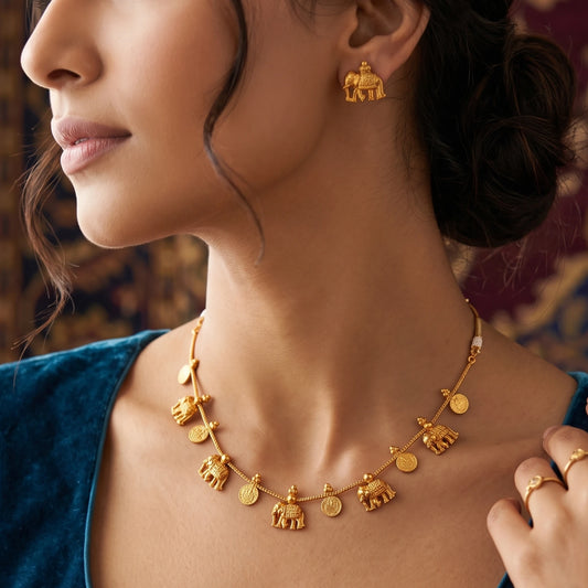A close-up lifestyle shot of a model wearing a gold toned necklace and matching gold toned earring, both featuring elephant and coin charms, set against a warm, blurred background.