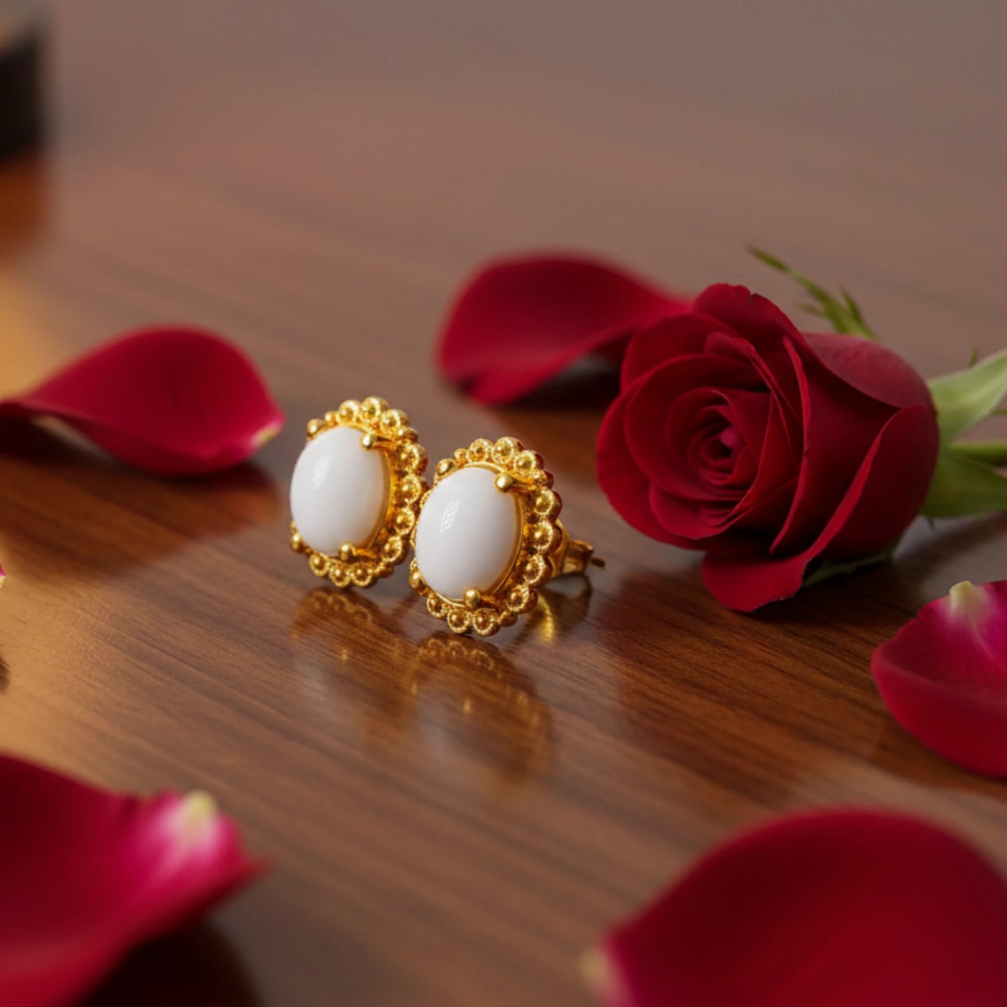 Gold plated earrings with oval white stones on a wooden surface with red roses and petals.