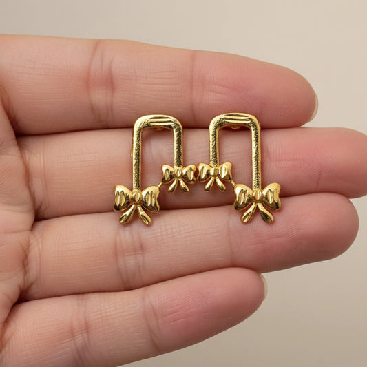 Gold plated ribbon bow-shaped earrings held between fingers against a neutral background