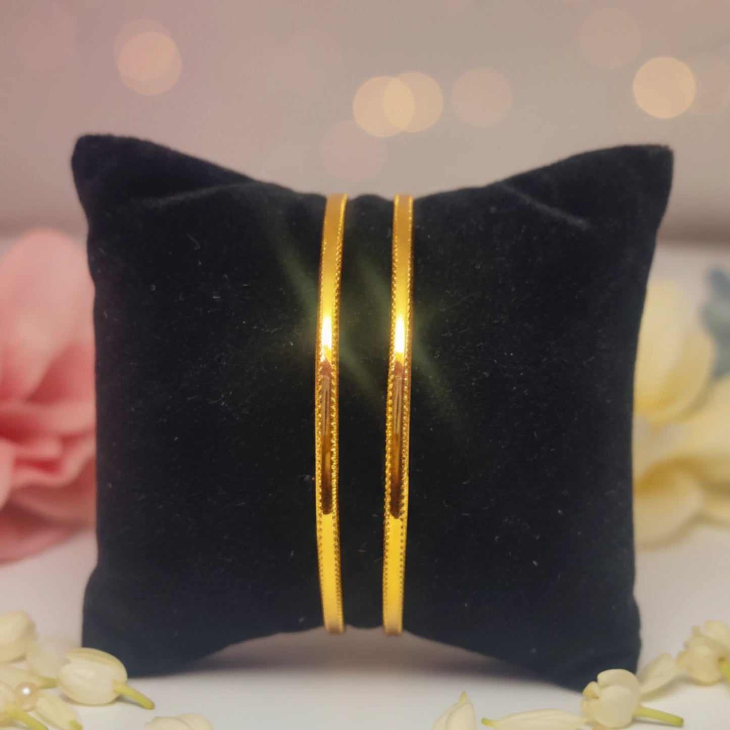 Two gold plated daily wear minimalist sleek bangles on a black pillow with a blurred floral background