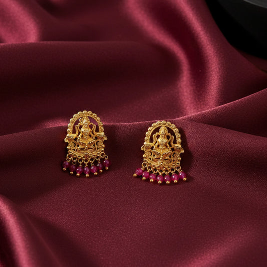 Gold plated earrings with lakshmi motif and ruby red gemstones on a maroon fabric background