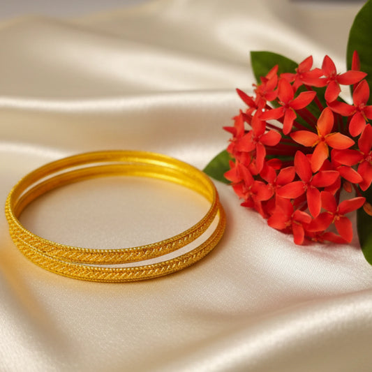 Two gold plated bangles with classic micro texturing on a beige fabric background with red flowers.