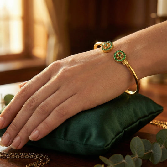 Hand wearing a gold plated bracelet with green gemstones on a green cushion.