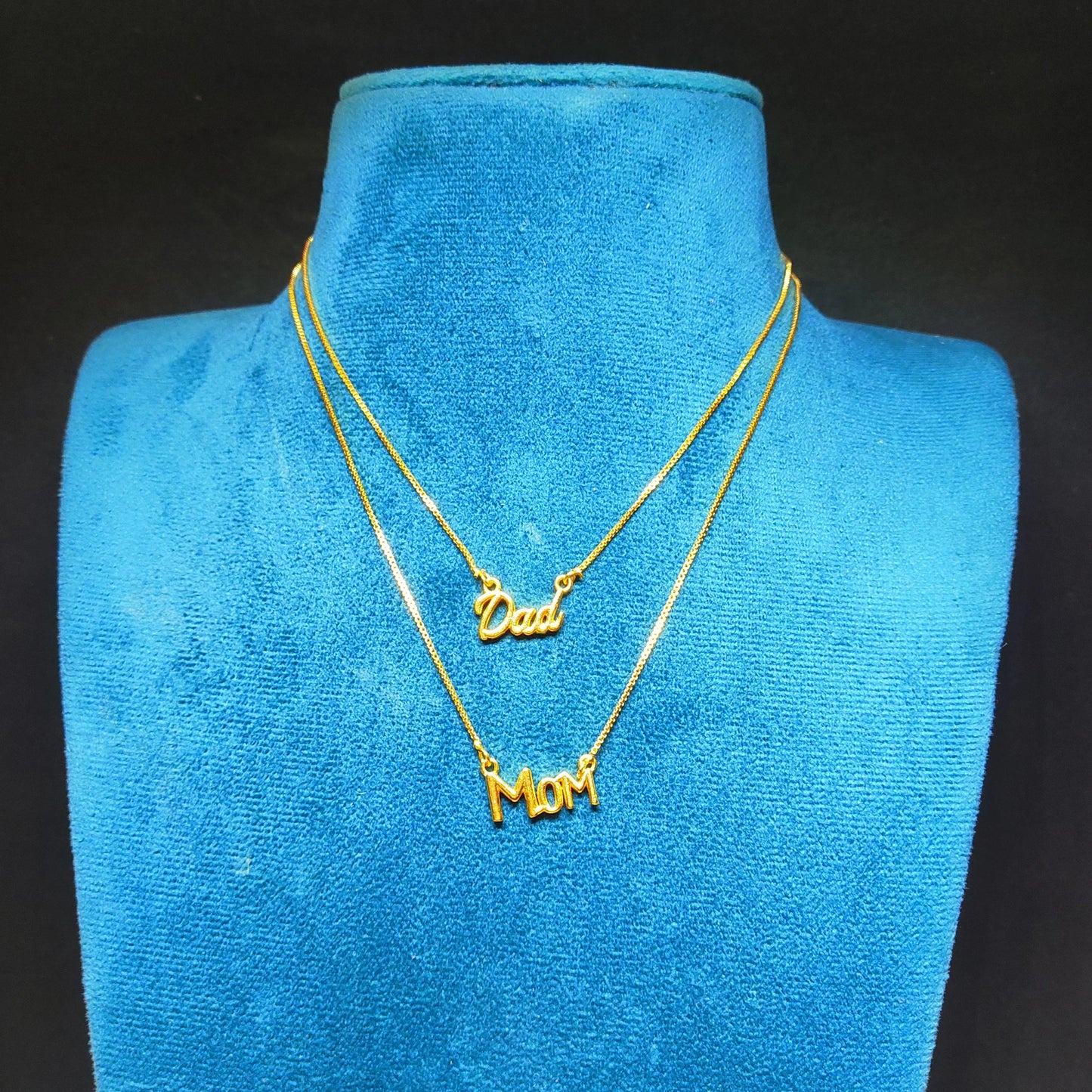 Gold plated neck chain with 'Dad' and 'Mom' pendants on a blue velvet stand.