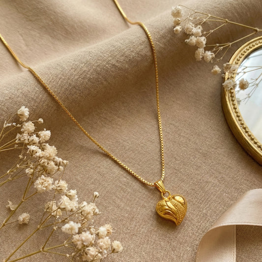 Gold plated neckchain with a heart pendant on a beige fabric background with dried flowers.