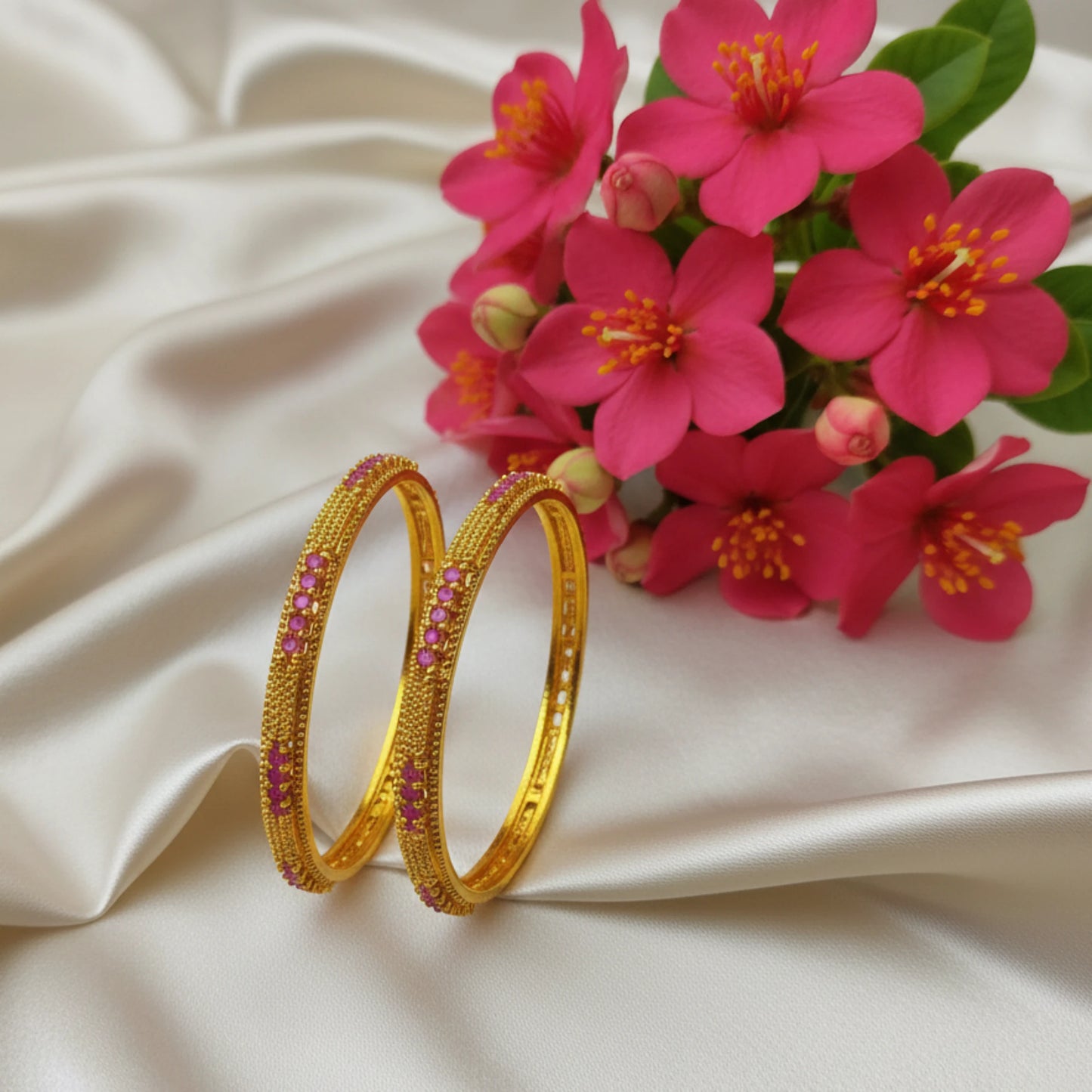 Gold plated bangles with pink stones and filigree work on a beige fabric background with pink flowers.