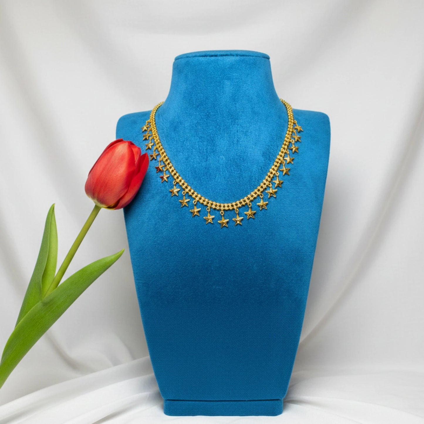 Gold plated necklace with star charms or pendants on a blue velvet mannequin bust with a red tulip, against a white background