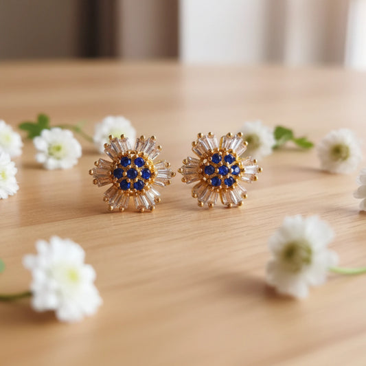 Gold plated floral stud earrings with blue and clear stones on a wooden surface with white flowers.