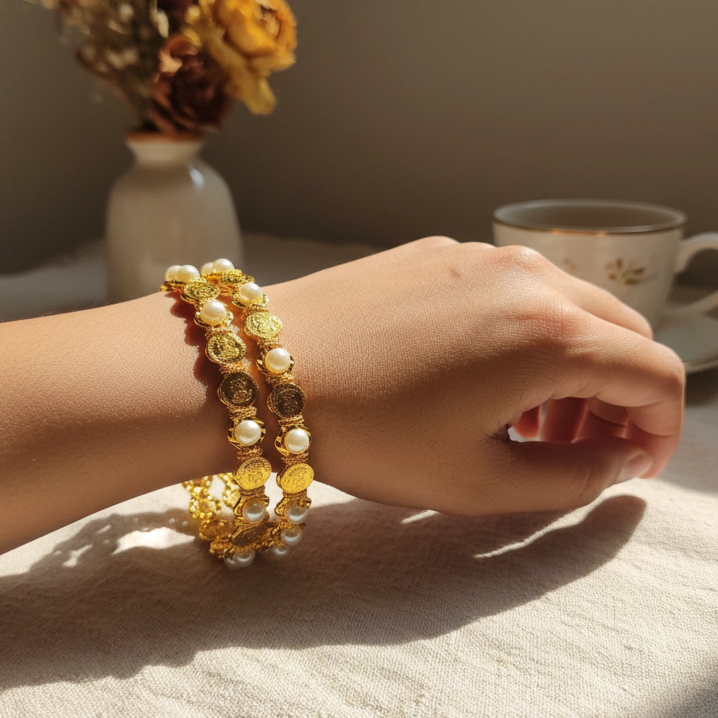 Hand wearing gold plated bangles with lakshmi motif and pearl in a intricate design with a cup of tea and flowers in the background