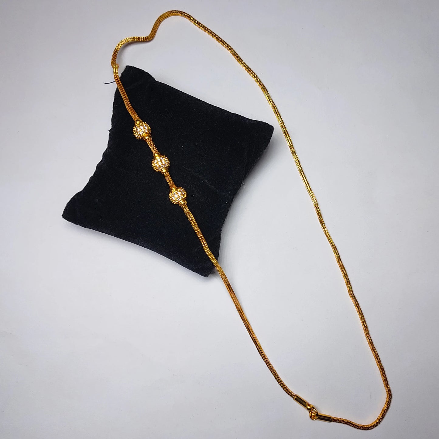 Gold plated mugappu neckchain with white stones on a black cushion against a light gray background