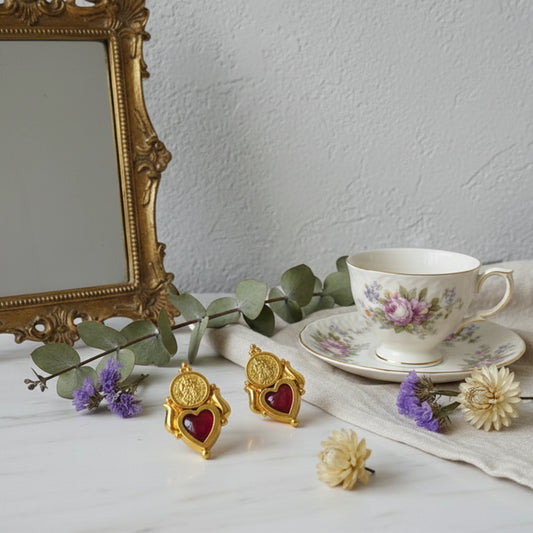 Gold plated heart-shaped palakka earrings with ruby red inlays on a table with a teacup, saucer, and flowers.