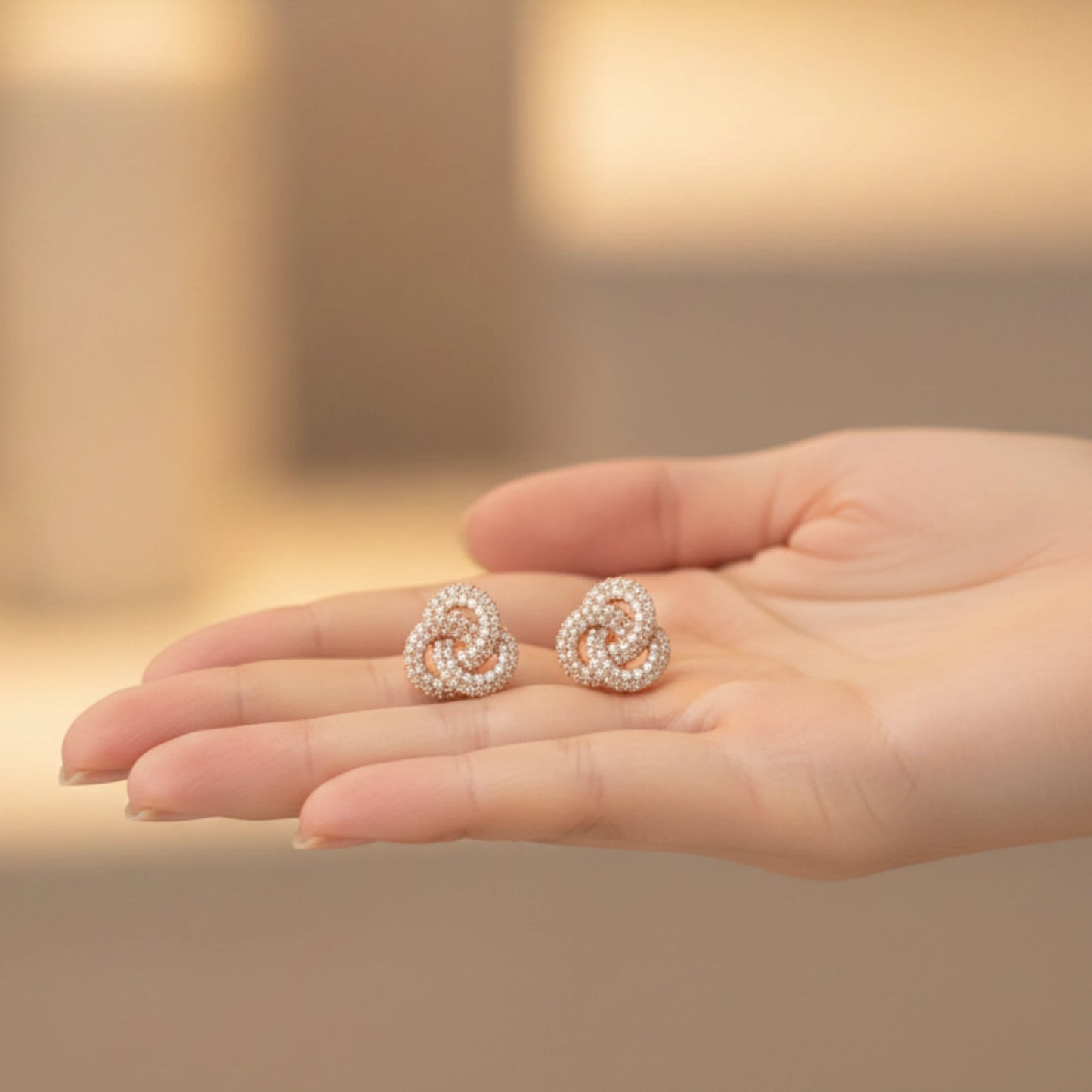 Pair of rose gold earrings with rose knot design held in a hand with a blurred background