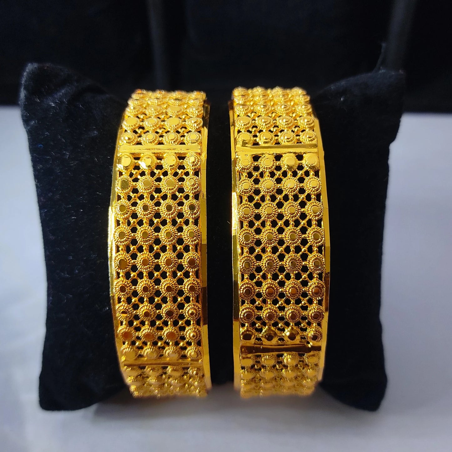 Pair of gold-plated bangles with Jali work and intricate designs on a black cushion.