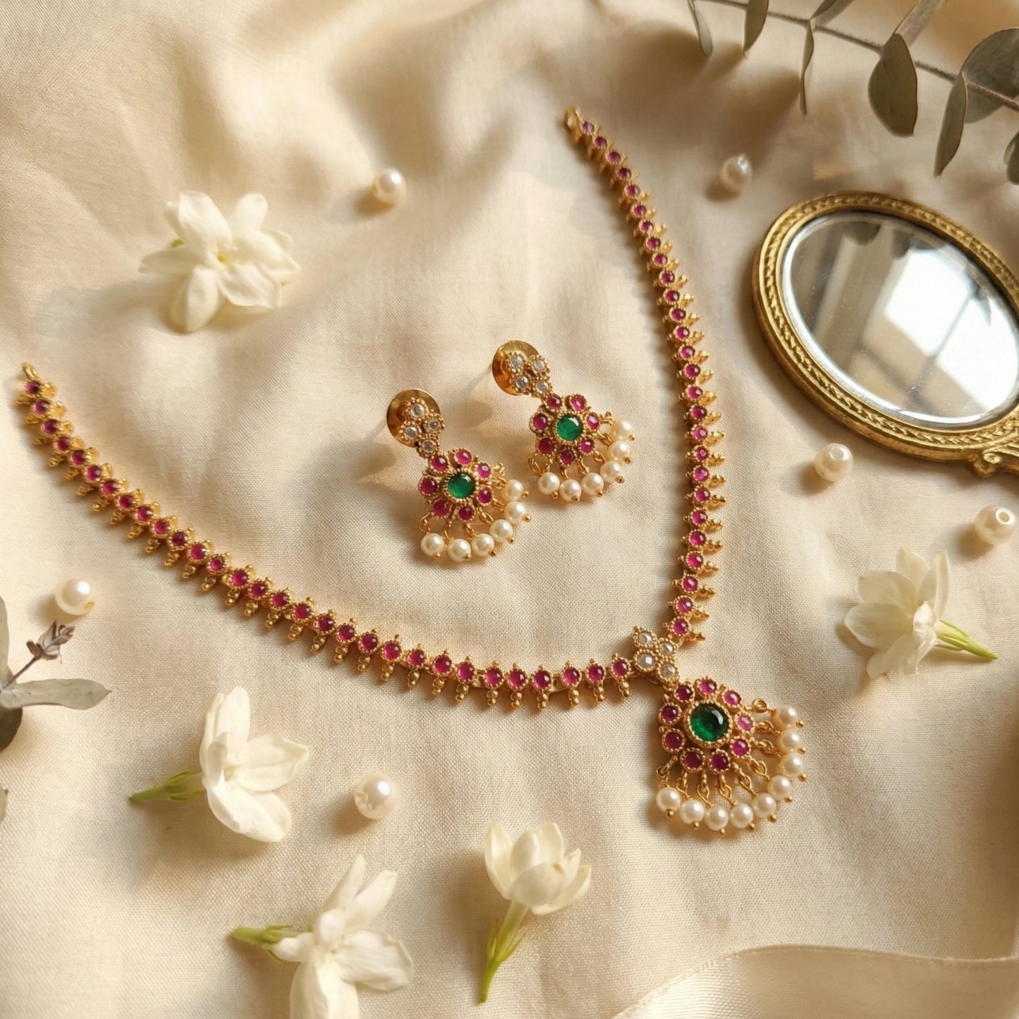 Gold toned jewelry set with pearls and gemstones on a soft fabric background with flowers and a mirror.