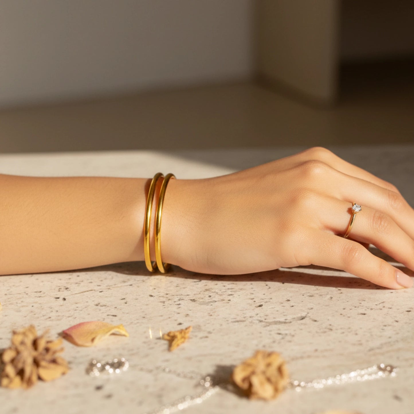 Hand wearing gold plated minimalist stackable bangles with a ring, on a textured surface with dried flowers.