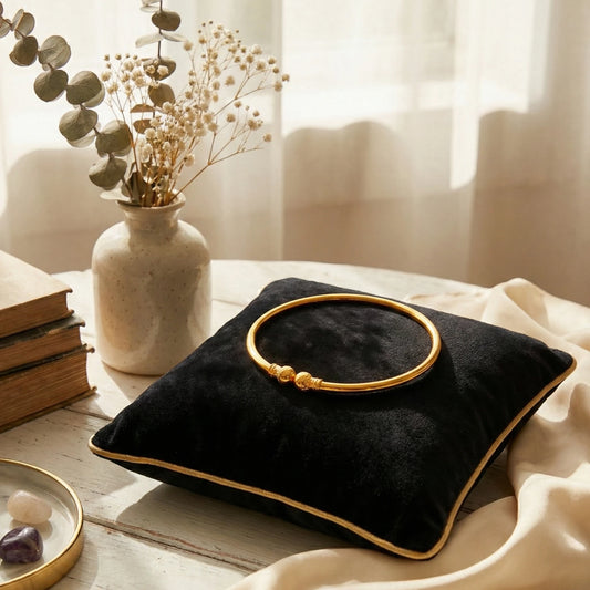Black pillow with gold plated Kada on a table next to a vase with flowers and books.