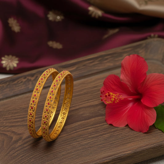 Gold plated bangles with ruby red stones on a wooden surface next to a red flower