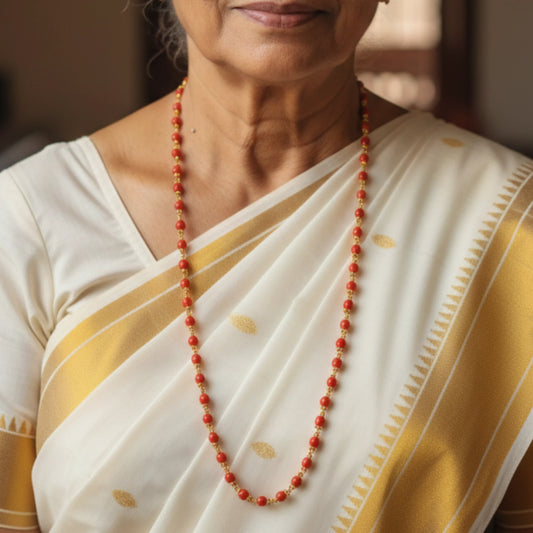 Woman wearing a white saree with gold border and a red pavizham beaded necklace.