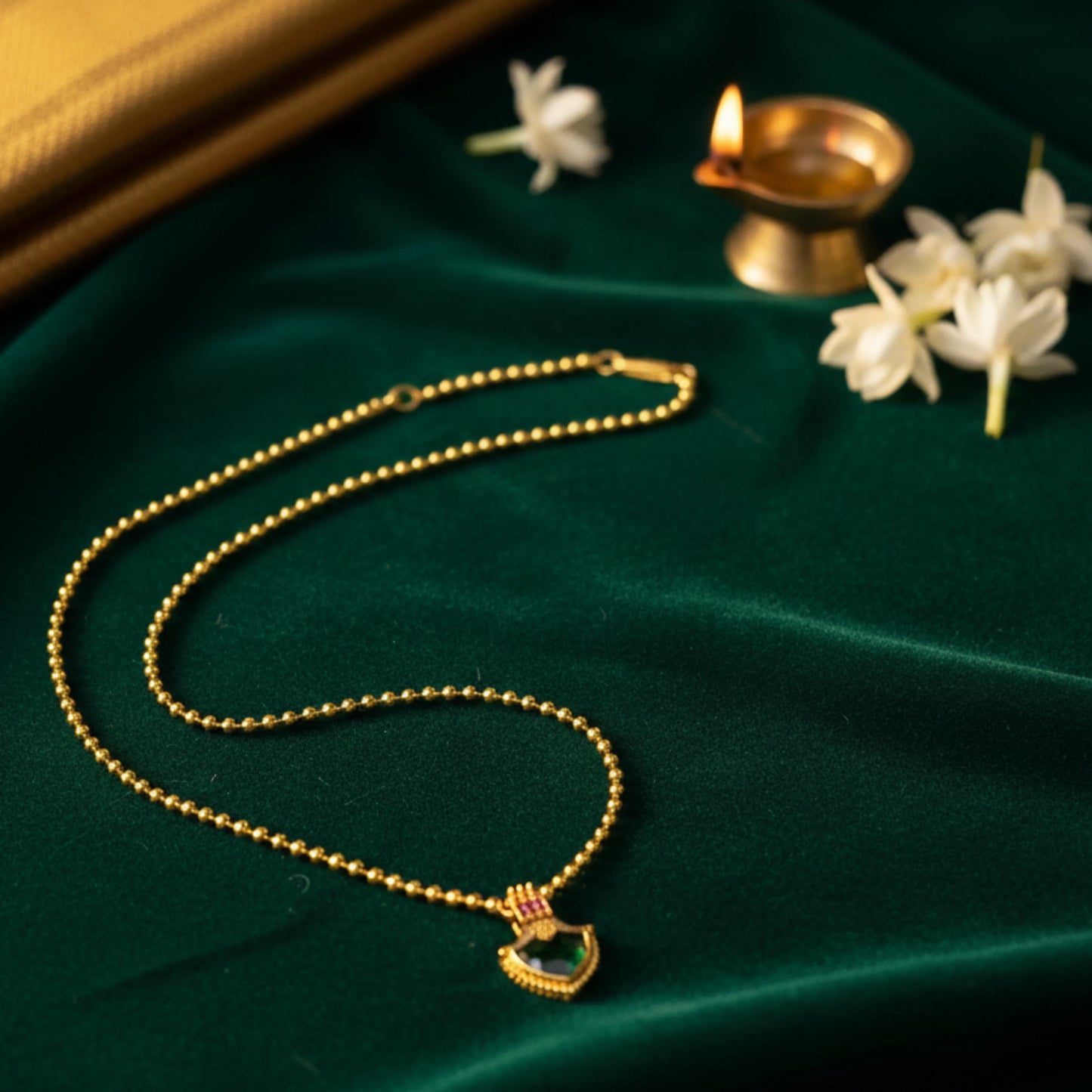 Gold plated necklace with a heart palakka pendant on a green fabric background with flowers and a lamp.