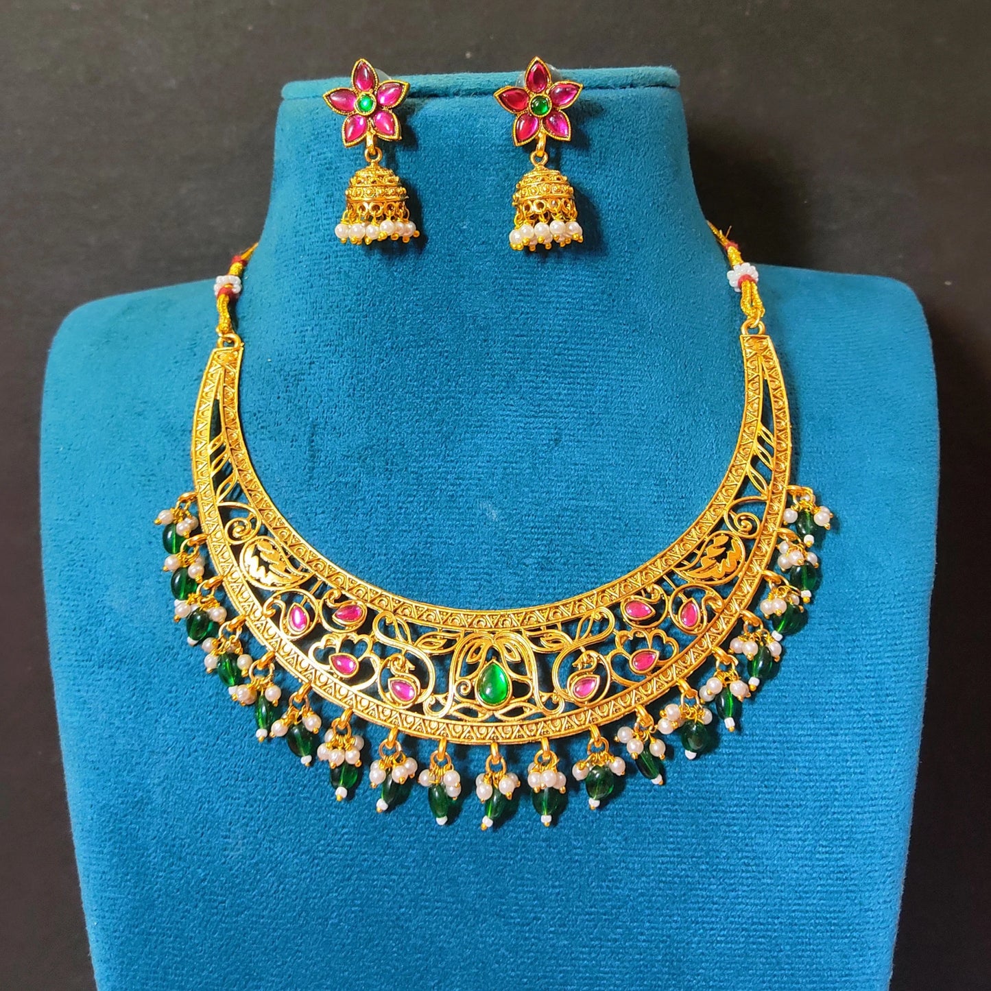 Antique gold plated crescent Hasli choker necklace set with intricate openwork, green bead drops, pink stone accents, and matching flower-shaped jhumka earrings on a blue display stand.