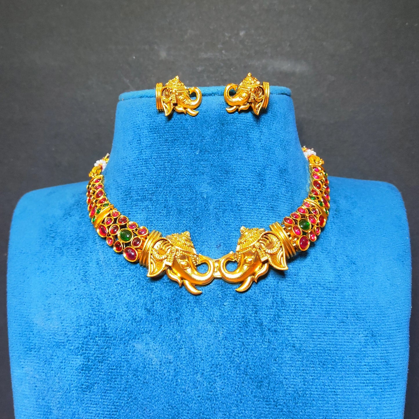 Gold-toned jewelry set displayed on a blue velvet mannequin bust against a dark gray background. The set includes a choker necklace with elephant and floral motifs, matching stud earrings, and a small box.