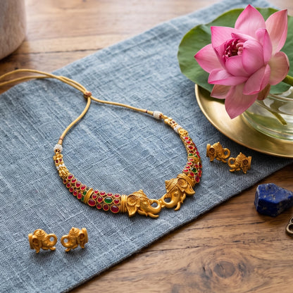 Gold-toned jewelry set featuring a necklace with elephant motifs and red and green bead accents, accompanied by matching earrings, arranged on a blue fabric cloth over a wooden surface with a pink lotus flower and a gold dish nearby.