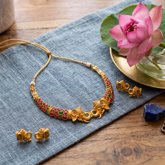 Gold-toned jewelry set featuring a necklace with elephant motifs and red and green bead accents, accompanied by matching earrings, arranged on a blue fabric cloth over a wooden surface with a pink lotus flower and a gold dish nearby.
