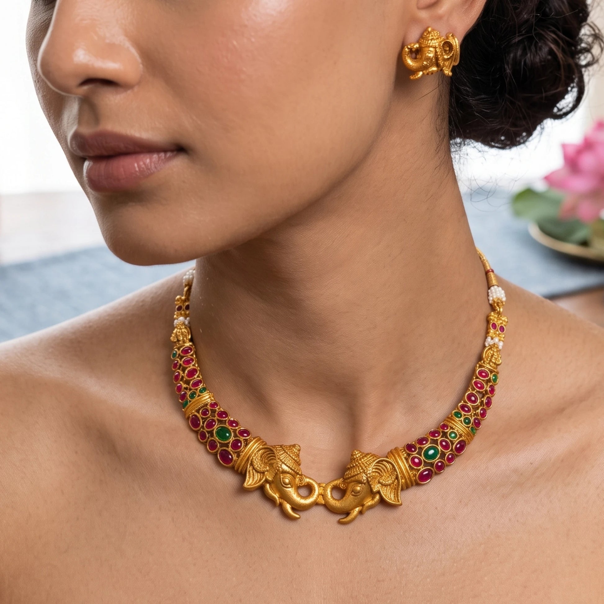 A close-up lifestyle shot of a model wearing a gold-toned Ganesha necklace and matching Ganesha earrings. The necklace features detailed Ganesha/elephant/gajah figures and is embellished with red and green stones, set against a softly blurred background.