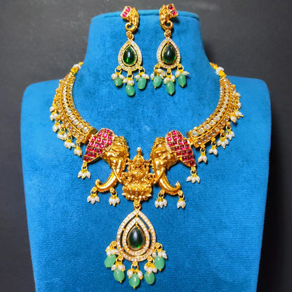 Antique Gold Finish Indian traditional jewelry set featuring a necklace which has Goddess Lakshmi flanked with twin elephants and matching earrings with green teardrop stones, white pearl-like accents, displayed on a blue velvet stand.