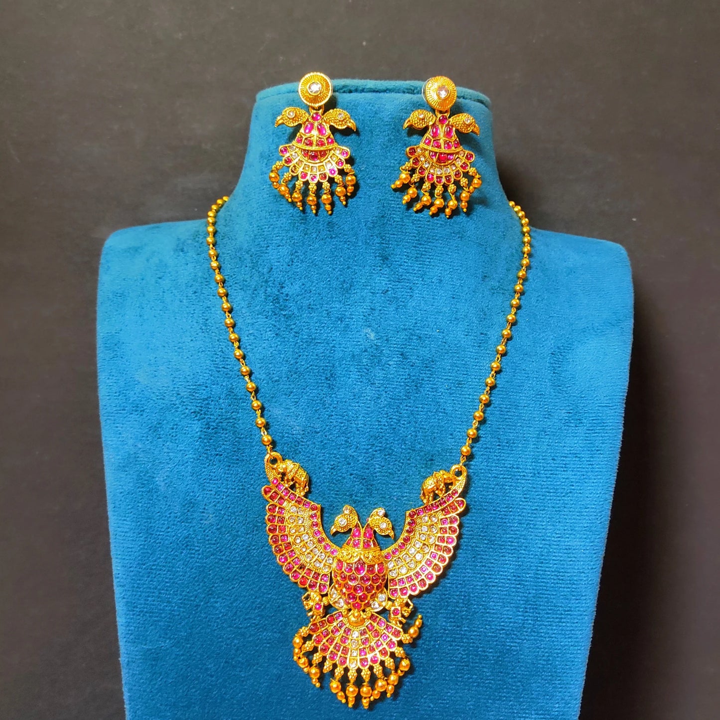 A traditional South Indian Gandaberunda temple jewelry set featuring a matte antique gold toned pendant necklace and matching earrings. The intricate pendant depicts the mythical royal two-headed bird adorned with radiant ruby-red kemp stones and delicate dangling gold toned beads, suspended from a gold toned bead chain.