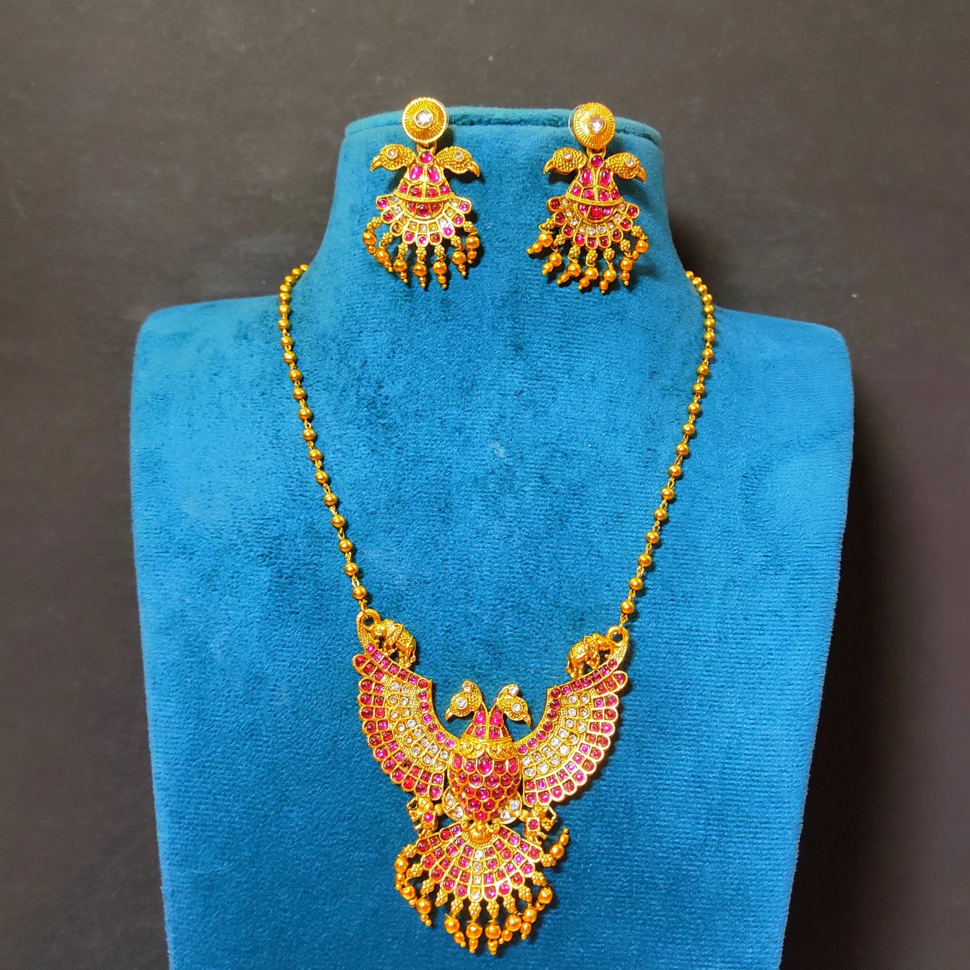 A traditional South Indian Gandaberunda temple jewelry set featuring a matte antique gold toned pendant necklace and matching earrings. The intricate pendant depicts the mythical royal two-headed bird adorned with radiant ruby-red kemp stones and delicate dangling gold toned beads, suspended from a gold toned bead chain.