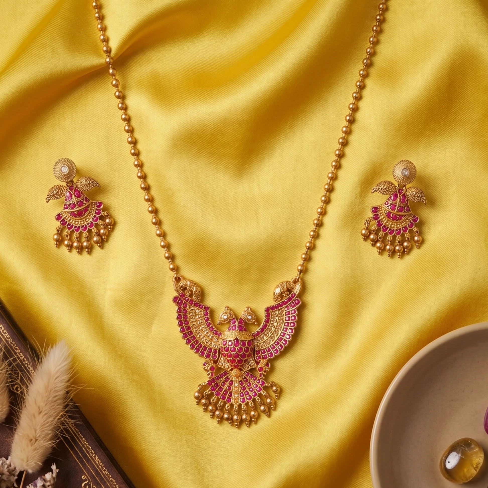 A traditional South Indian Gandaberunda temple jewelry set featuring a matte antique gold toned pendant necklace and matching earrings. The intricate pendant depicts the mythical royal two-headed bird adorned with radiant ruby-red kemp stones and delicate dangling gold toned beads, suspended from a gold toned bead chain.