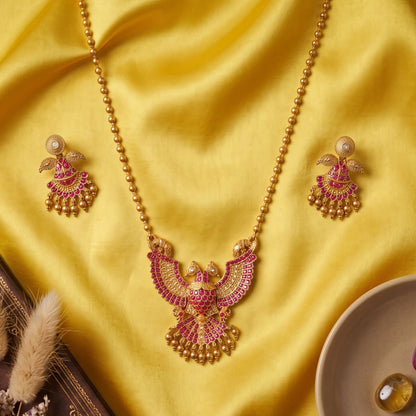A traditional South Indian Gandaberunda temple jewelry set featuring a matte antique gold toned pendant necklace and matching earrings. The intricate pendant depicts the mythical royal two-headed bird adorned with radiant ruby-red kemp stones and delicate dangling gold toned beads, suspended from a gold toned bead chain.