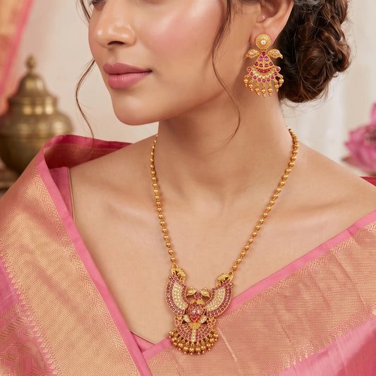 A traditional South Indian Gandaberunda temple jewelry set featuring a matte antique gold toned pendant necklace and matching earrings. The intricate pendant depicts the mythical royal two-headed bird adorned with radiant ruby-red kemp stones and delicate dangling gold toned beads, suspended from a gold toned bead chain.
