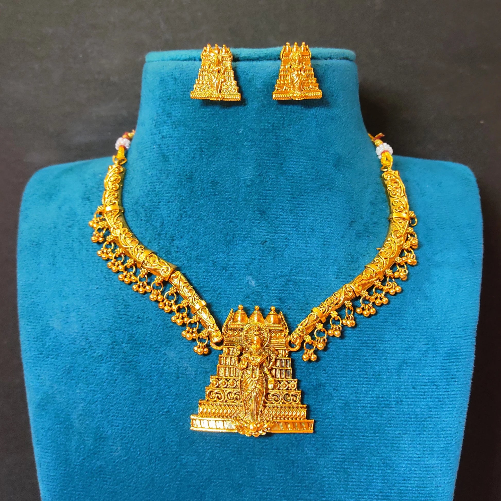 The necklace features a curved, rigid gold toned collar band adorned with a delicate fringe of dangling micro-gold beads, anchored by a prominent, tiered temple gopuram tower pendant intricately engraved with a divine deity figure.