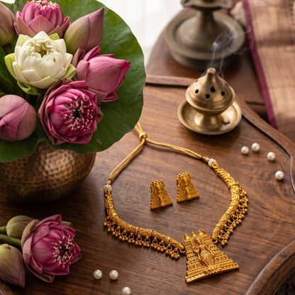 The necklace features a curved, rigid gold toned collar band adorned with a delicate fringe of dangling micro-gold beads, anchored by a prominent, tiered temple gopuram tower pendant intricately engraved with a divine deity figure.
