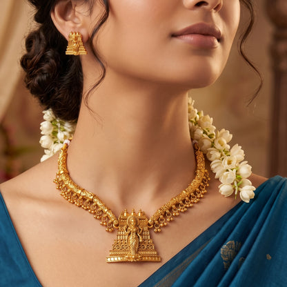 The necklace features a curved, rigid gold toned collar band adorned with a delicate fringe of dangling micro-gold beads, anchored by a prominent, tiered temple gopuram tower pendant intricately engraved with a divine deity figure.