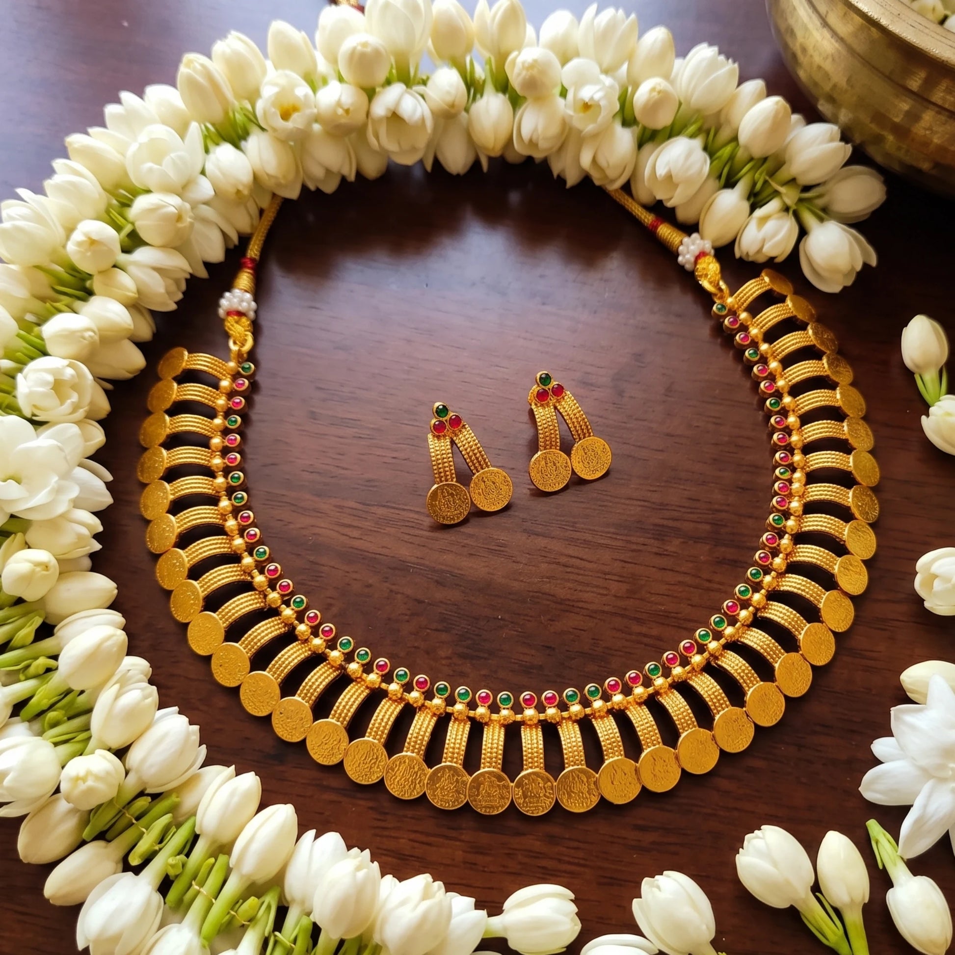 Antique gold finish coin-style necklace and matching earrings displayed on a dark wooden surface, surrounded by white flower garlands. The necklace features rows of gold toned coins with small red and green bead accents, and the earrings are gold toned with red and green stone details.