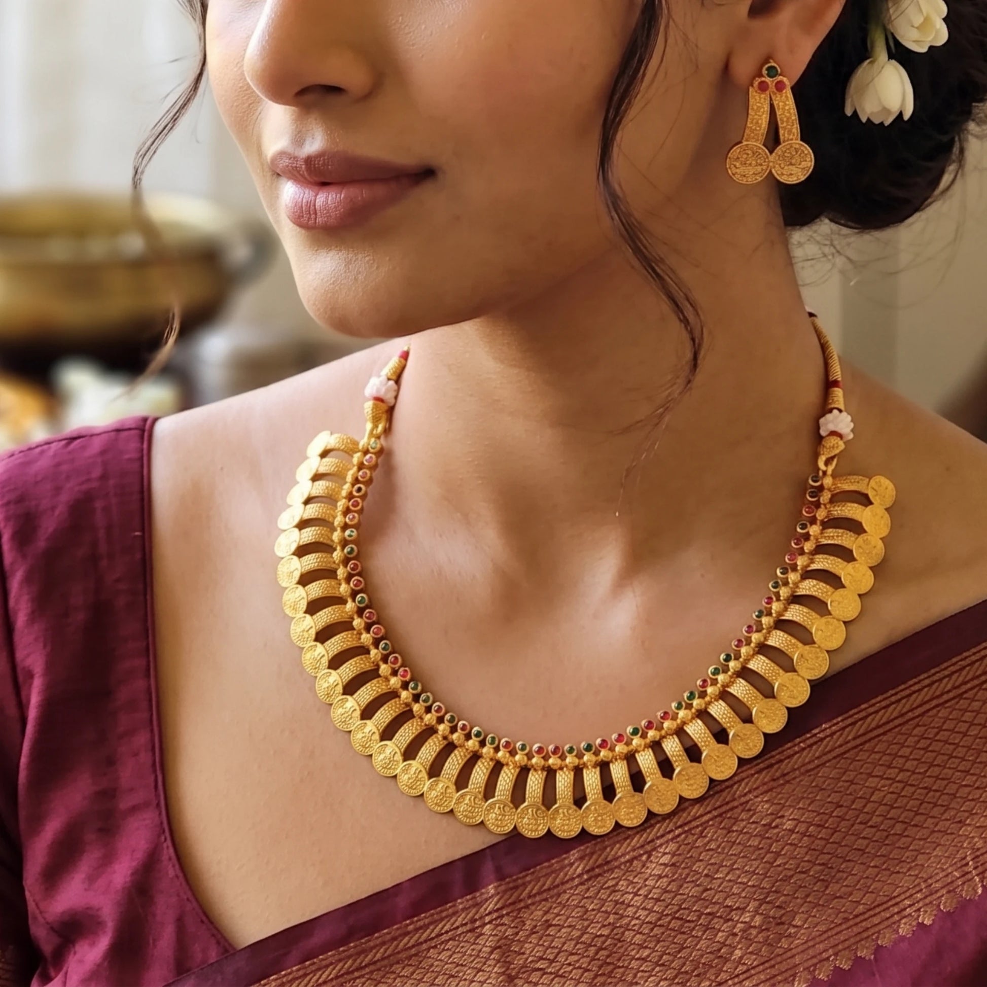 A close-up of a woman wearing a gold-toned traditional collar necklace with scalloped coin-like edges and small red and green bead accents, along with matching dangling earrings.