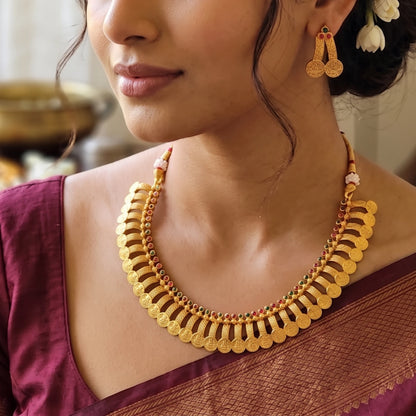 A close-up of a woman wearing a gold-toned traditional collar necklace with scalloped coin-like edges and small red and green bead accents, along with matching dangling earrings.
