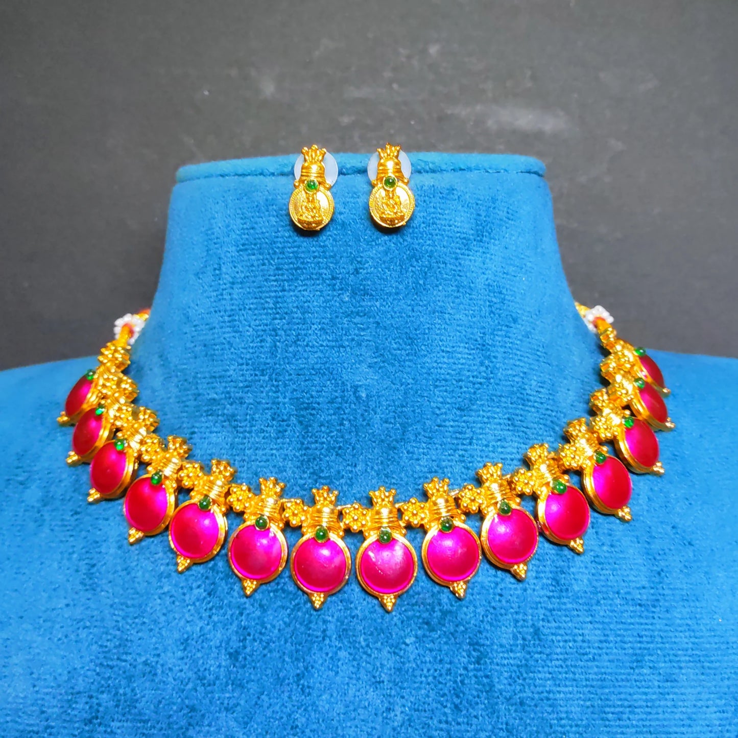 Antique Gold-toned traditional Indian jewelry set featuring a necklace and matching earrings with bright pink enamel coated pendants and small green accent stones, displayed on a blue velvet stand against a dark gray background.