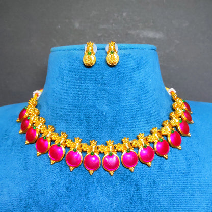 Antique Gold-toned traditional Indian jewelry set featuring a necklace and matching earrings with bright pink enamel coated pendants and small green accent stones, displayed on a blue velvet stand against a dark gray background.