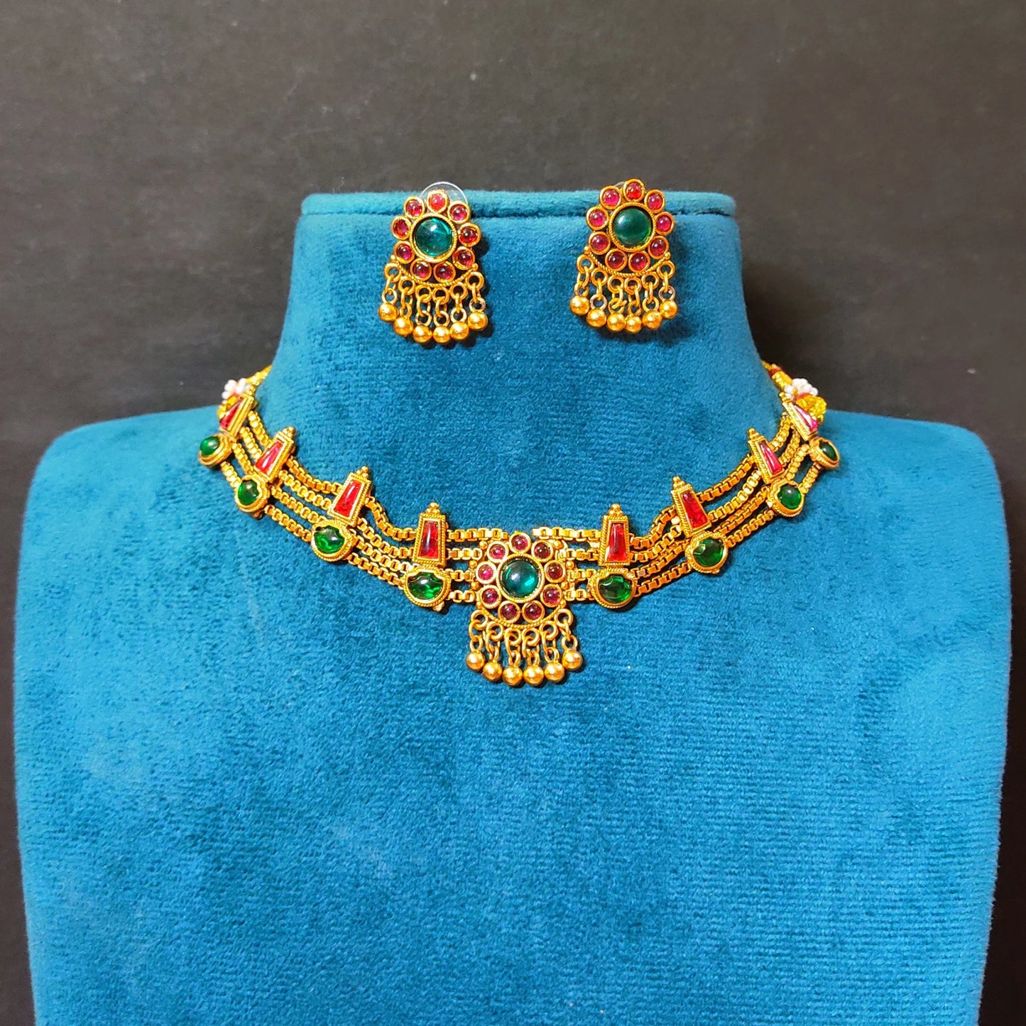 A traditional South Indian multi-strand choker necklace and matching stud earrings in a matte antique gold finish, displayed on a blue velvet jewelry bust against a dark gray background. The necklace features triple-layered delicate gold chains accented with alternating green and red kemp stones, centered around a circular floral medallion with dangling gold beads.