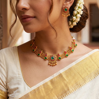 A traditional South Indian multi-strand choker necklace and matching stud earrings in a matte antique gold finish, displayed on model. The necklace features triple-layered delicate gold chains accented with alternating green and red kemp stones, centered around a circular floral medallion with dangling gold beads.